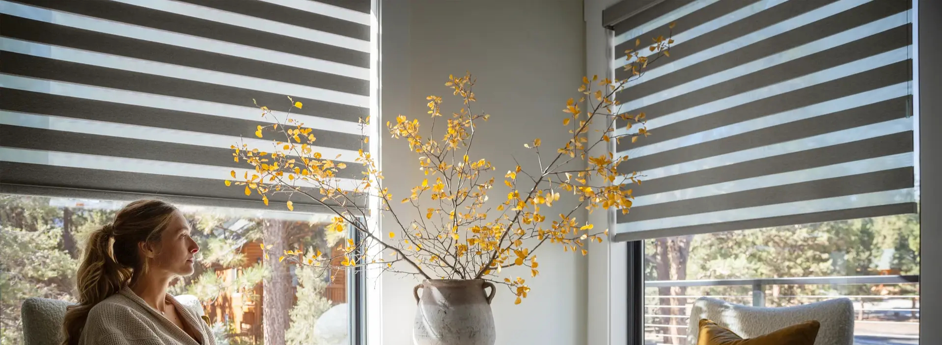 Woman relaxing in a sunlit living room with gray zebra shades on corner windows and a vase of golden autumn branches
